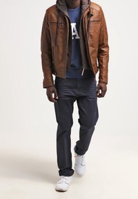 Veste en cuir marron avec une couche à capuche, sweat-shirt bleu marine avec texte blanc, pantalon foncé et baskets blanches. Design simple et moderne.