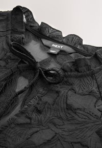 Top nero trasparente con ricami floreali, scollo arricciato e dettaglio a fiocco sul davanti. Etichetta con la scritta "NEXT." Tessuto strutturato con design intricato.