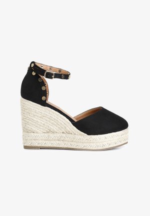 Black espadrille wedge shoe with ankle strap, gold studs, and braided jute platform sole on white background.
