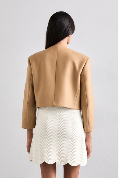 Veste courte couleur camel avec des épaules structurées, associée à une robe blanche à texture ondulée. Tissu lisse avec un minimum de détails.