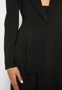 Černý tailored sak s hlubokým výstřihem do V, zpevněnými rameny a peplum střihem. Hladká tkanina s viditelnými švy.