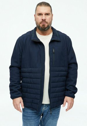 Man met baard die een marineblauwe doorgestikte jas draagt over een wit overhemd en blauwe jeans, staand tegen een effen witte achtergrond.