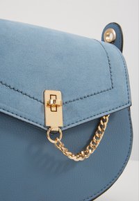 Borsa a mano in pelle blu con texture scamosciata, chiusura in metallo dorato e dettaglio a catena. Presenta una chiusura a patta con un design cucito.