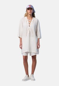 Tunic in lino con scollatura a lacci, maniche lunghe e vestibilità comoda; abbinata a pantaloni corti bianchi e sneaker; include un cappello con disegno.