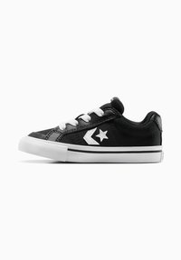 Zapatillas de lona negras con suela y cordones de goma blanca, que presentan un logo de estrella plateada y un diseño de chevrón en el lateral. Silueta baja.