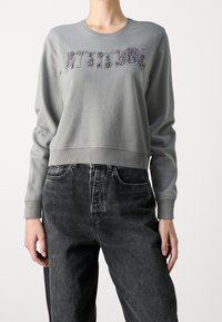 Sudadera gris corta con letras texturizadas "ACTITUD", cuello redondo y puños acanalados, combinada con vaqueros negros de cintura alta.