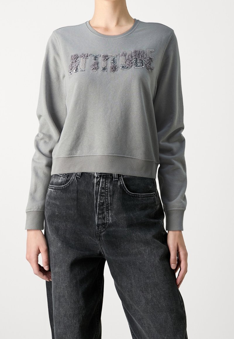 Sudadera gris corta con letras texturizadas "ACTITUD", cuello redondo y puños acanalados, combinada con vaqueros negros de cintura alta.