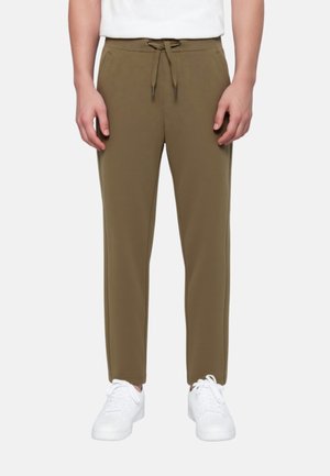 Mann trägt olivgrüne Jogginghose mit Kordelzug in der Taille und weiße Sneakers, stehend vor einem einfarbigen Hintergrund.