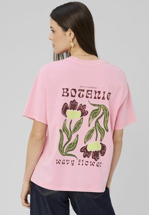 Femme aux longs cheveux noirs portant un T-shirt rose avec des illustrations florales vertes et brunes et le texte "Botanic wavy flower" au dos.