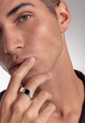 Nahaufnahme eines Mannes, der seine Lippen mit Zeige- und Mittelfinger berührt und dabei einen breiten silbernen Ring am Mittelfinger trägt.