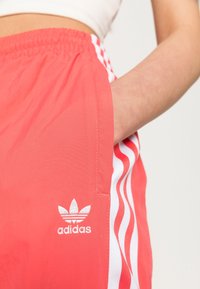 Detailní záběr osoby v korálových šortkách Adidas s bílými pruhy a logem, ruka pohodlně spočívající v kapse.