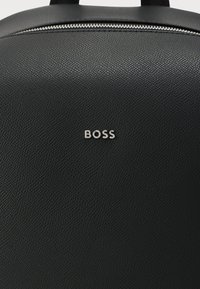 Mochila negra con material texturizado, prominente logotipo plateado "BOSS" y cierre con cremallera en la parte superior.