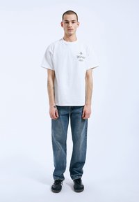 Bílá bavlněná tričko s krátkým rukávem a minimální grafikou na levé straně hrudi, v kombinaci s modrými rovnými džíny a černými teniskami.