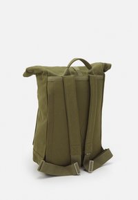 Olive green canvas backpack with roll-top closure, adjustable straps, and a top handle. Durable texture and minimalist design with no visible logos.