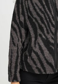 Veste en polaire grise et noire avec un motif zèbre texturé, dotée d'une fermeture éclair à l'avant et de manches longues. Tissu doux et chaud.