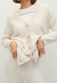 Gebreid crème vest met een getextureerd patroon, v-hals en parelknopen. Gecombineerd met bijpassende crème getextureerde broek.