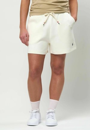 Inimene, kes kannab kreemika värvi nööriga lühikesi pükse, beeži värvi topi, heleroosa sokke ja valgeid vabaajajalatsid, seisab ühevärvilise tausta ees.