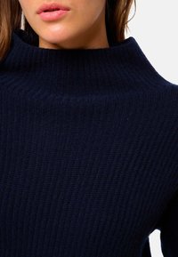 Pull en mailles côtelées bleu marine avec un col haut et large. Le tissu semble doux et texturé, mettant en valeur des côtes verticales tout au long.