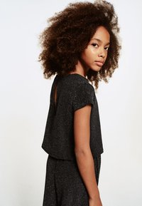 Svart glittrig topp med korta ärmar och en nyckelhålsdetalj i ryggen. Mjuk textur och lös passform som skapar en avslappnad silhuett.