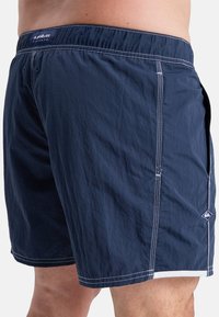 Navy blue swim shorts in lightweight fabric with white stitching. Features an elastic waistband and side pockets, with a logo on the hem.