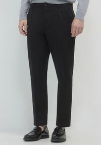 Schwarze maßgeschneiderte Hosen aus glattem Stoff, mit flacher Vorderseite, seitlichen Taschen und Knopfverschluss. Kombiniert mit glänzenden schwarzen Schuhen.