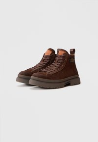 Zapatillas altas de ante marrón con suela de goma gruesa, que presentan un diseño de cordones, logotipo en relieve y una lengüeta reforzada en el talón para facilitarn su uso.