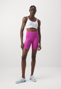 Bijela sportska bralette s trakama s logotipom, uparena s jarko ružičastim kratkim hlačama visokog struka. Model nosi bijele i plave tenisice.