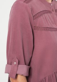 Długa bluzka w kolorze wrzosowym z długimi rękawami, plisami, koronkowymi akcentami i zapinaniem na guziki przy szyi. Rękawy podwinięte z guzikiem. Teksturowana tkanina.