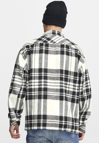 Camicia di flanella a quadri bianco e nero con un fit rilassato, maniche lunghe e colletto tradizionale, abbinata a jeans in denim e un berretto.