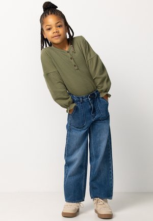 Olijfgroene top met lange mouwen van textuurstof en knopen, gecombineerd met wijde blauwe denim jeans. Model draagt beige sneakers.