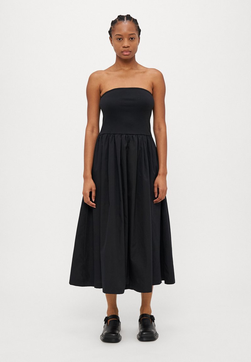 Svart off-shoulder kjole med tettsittende liv og voluminøs, rynket skjørt. Glatt, lett stoff med matt overflate.