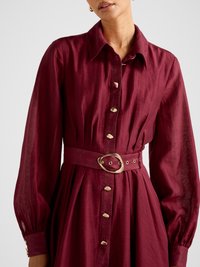 Žena v bordových šatách s dlhými rukávmi z textúrovaného materiálu, so zlatými nepravidelnými gombíkmi a zodpovedajúcim opaskom so zlatou prackou, zobrazená od krku po boky.