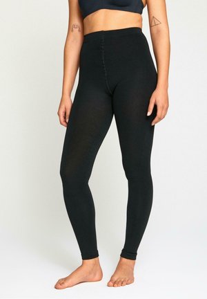 Nainen, jolla on mustat korkeavyötäröiset leggingsit ja tumma urheiluliivit, molemmissa käsivarsissa minimaalinen määrä tatuointeja, seisoo paljain jaloin yksinkertaisella taustalla.