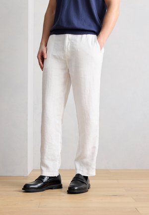 Osoba oblečená v bielych ľanových nohaviciach, čiernych kožených poltopánkach a tmavomodrom tričku, stojaca na drevenej podlahe pri šedej stene.