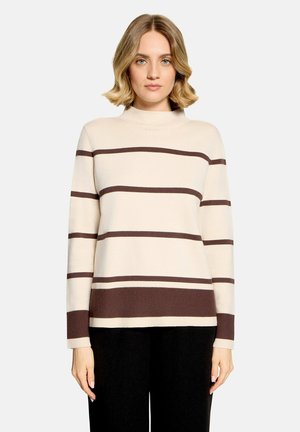 Femme aux cheveux blonds portant un pull crème avec des rayures horizontales marron et un pantalon noir sur un fond clair uni.