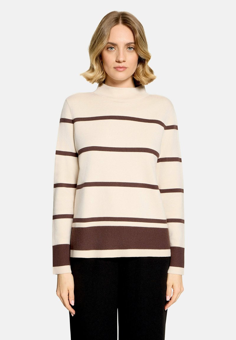 Femme aux cheveux blonds portant un pull crème avec des rayures horizontales marron et un pantalon noir sur un fond clair uni.
