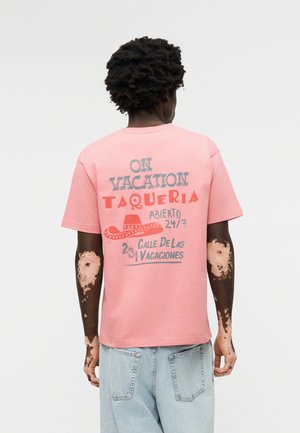 Personne aux cheveux bouclés foncés et atteinte de vitiligo sur les bras porte un T-shirt rose avec le texte « On Vacation Taqueria » et un graphique de sombrero dans le dos.