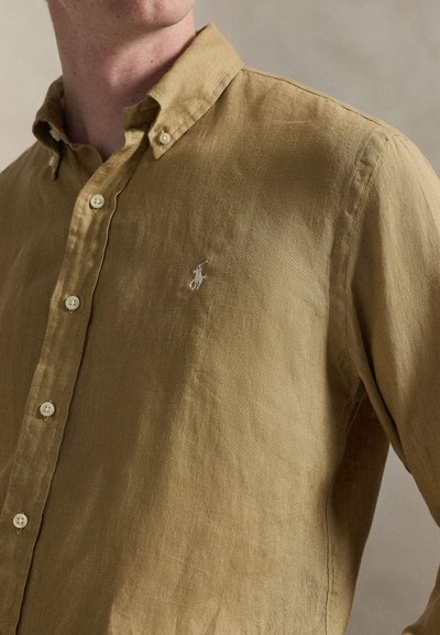 Jasnobrązowa lniana koszula z kołnierzykiem na guziki i małym białym logo na piersi. Posiada białe guziki i fakturowane wykończenie.