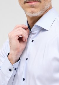 Camicia bianca con sottili righe blu, dotata di un colletto interno a pois scuri e bottoni scuri. Tessuto texture visibile.