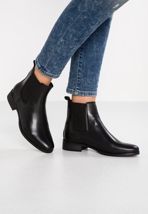 Botas de cuero negro hasta el tobillo con punta afilada, paneles laterales elásticos y un tacón bajo en bloque, combinadas con unos vaqueros ajustados azules.