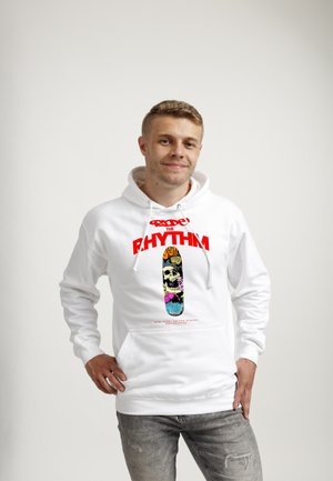 Młody mężczyzna w białej bluzie z kapturem z kolorową grafiką deskorolki i napisem „Ride the Rhythm”, stojący z ręką na biodrze na jednolitym tle.