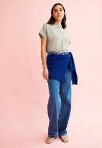 Lichtgroen shirt met korte mouwen, blauwe wikkelrok met knoopdetails, wijde blauwe jeans, en beige instapschoenen tegen een roze achtergrond.