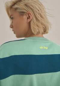 Zelený a námořnický pruhovaný svetr s kulatým výstřihem a krátkým rukávem. Na zadní straně je žlutý vyšitý nápis "oh hey". Měkká textura.