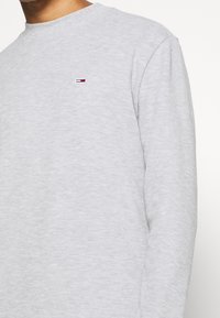 Grauer Langarm-Sweatshirt mit Rundhalsausschnitt, weichem Material und einem kleinen gestickten Logo in Rot, Weiß und Blau auf der Brust.