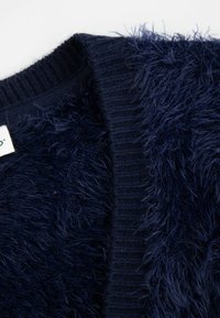 Granatowy kardigan z miękką, puszystą teksturą z jednej strony i prążkowanym splotem z drugiej, z widoczną naszywką.