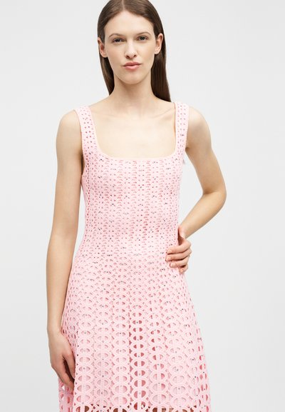 Jeune femme aux cheveux bruns raides portant une robe en crochet rose sans manches, debout avec une main sur la hanche sur un fond blanc.