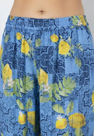 Mellemsektion af person iført blå bukser med gult citron- og grønt bladmønster, elastisk taljebånd synligt mod lys baggrund.
