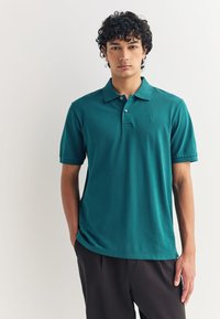 Turkio spalvos polo marškinėliai, pagaminti iš lygaus audinio, su apykakle, trijų mygtukų užsegimu ir subtiliu logotipu ant krūtinės. Trumpomis rankovėmis.