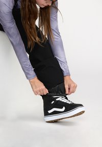 Baskets montantes noires avec des accents et des lacets blancs. Le porteur ajuste la chaussure tout en portant un haut à manches longues violet et un pantalon noir.