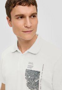 Weißes Poloshirt aus strukturiertem Stoff, mit einem grafischen Druck in Schwarz auf der linken Seite. Das Design umfasst Text und abstrakte Muster.